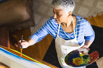 A mature biracial woman paints on canvas at home