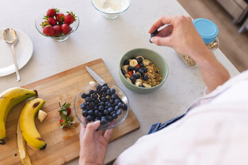 A person is preparing a healthy breakfast, adding blueberries to a bowl of granola and fruit
