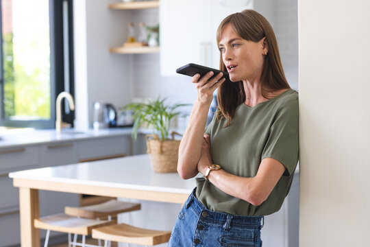 A middle-aged Caucasian woman is using voice command on her smartphone in a modern kitchen - Powered by Adobe