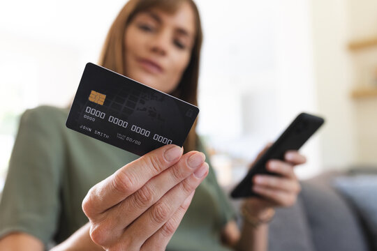 A middle-aged Caucasian woman is holding a credit card and a smartphone, managing her finances onlin