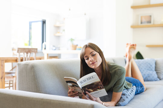 A young Caucasian woman relaxes on a couch while engrossed in a book, with copy space unaltered