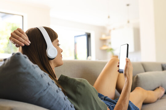 A young woman relaxes on a sofa with headphones and smartphone, with copy space unaltered