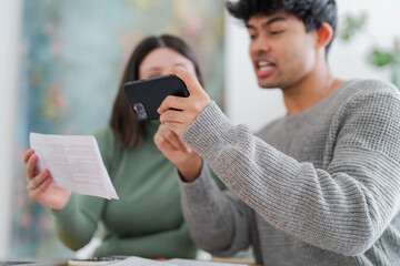 A young man captures an image of a paper document using his smartphone, with a focused expression as a woman looks on. The scene suggests a moment of sharing information or digitizing paperwork