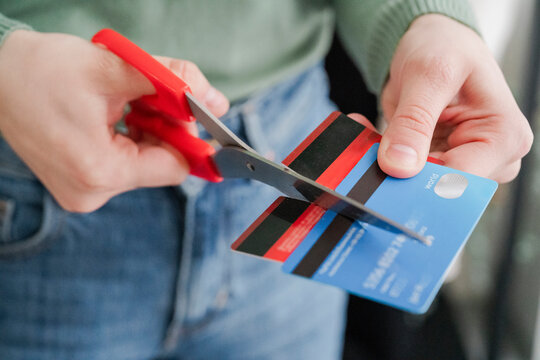 A person in a green sweater is seen cutting a stripe-patterned credit card with red scissors. The act signifies the intention to reduce debt or prevent overspending