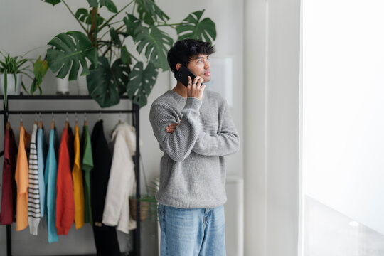 A young man stands by a window, lost in thought while talking on the phone. His casual attire and contemplative demeanor suggest a personal conversation