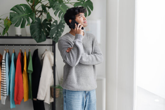 A young man stands by a window, lost in thought while talking on the phone. His casual attire and contemplative demeanor suggest a personal conversation