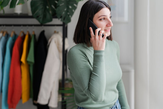 Smiling woman in a green sweater having a pleasant conversation on the phone, enjoying the comfort of her plant-adorned home