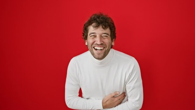 Joyful young hispanic man, standing in laughter wearing a sweater, belting out a hard laugh from a funny joke over a isolated red background