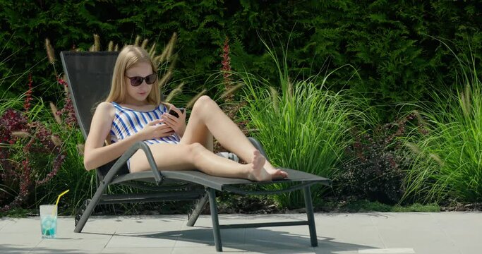 A teenager is relaxing on a sun lounger by the pool. Uses a smartphone
