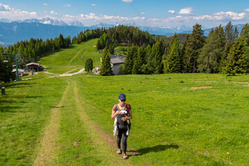 Woman with baby carrier on hiking trail along alpine meadow with scenic view of mountain range...
