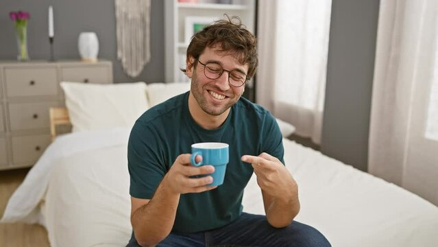 Cheerful young hispanic man, handsomely-bearded, joyfully pointing with confident gesture while sitting on bed, drinking coffee indoors, his optimistic smile revealing teeth in homey bedroom.