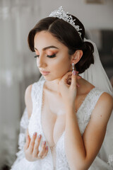 Beautiful young bride wearing earrings before wedding ceremony at home. A bride in a white wedding dress puts on earrings in her room in the morning.