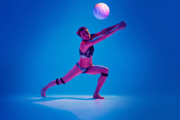 Fototapeta na wymiar Concentrated athlete woman, beach volleyball player passes ball from below against gradient blue background in pink neon light. Concept of sport, movement, active and healthy lifestyle, power.