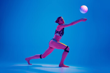 Fototapeta na wymiar Athlete woman in sportswear playing beach volleyball against gradient blue background in pink neon light. Concept of sport, movement, active and healthy lifestyle, power and strength.