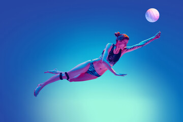 Athletic woman, volleyball player diving for ball in mid-air against gradient blue background in pink neon light. Concept of sport, movement, active and healthy lifestyle, power and strength.
