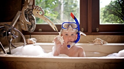 A young boy in the diving mask sits in the bath with the shell and listen to the sound from it 