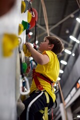 A young European boy in a yellow shirt is climbing a wall in an indoor climbing gym