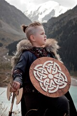A young boy in the viking cosplay costume holding a wooden shield and a axe and standing next to the beautiful alpine lake with Mountains view