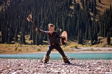 A young boy in the viking cosplay costume holding a wooden shield and a axe and training next to the beautiful alpine lake with Mountains view