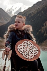 A young boy in the viking cosplay costume holding a wooden shield and a axe and standing next to the beautiful alpine lake with Mountains view
