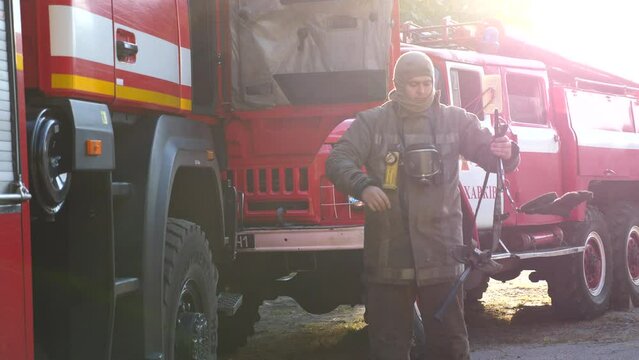 Male firefighter in balaclava fastening the belt on his uniform near a fire engine. Young fireman getting ready to goes to extinguishing fire against the background of a big red car. Slow motion