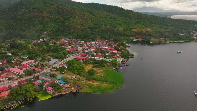 Traffic on coastal road at Taal Lake in Philippines. Green mountain with village and red roof houses. Aerial top down.