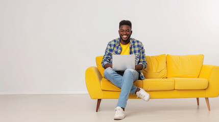 Happy young afro american man sitting in a yellow sofa and using a laptop on a white background. Networking, training, freelancing, remote work. - Powered by Adobe