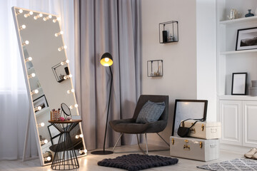 Makeup room. Stylish mirror with light bulbs, beauty products on table and armchair indoors