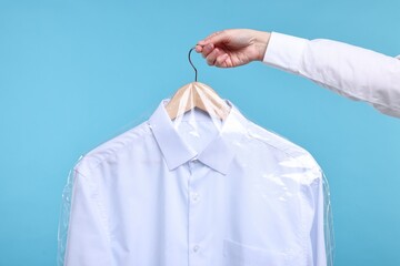 Dry-cleaning service. Woman holding shirt in plastic bag on light blue background, closeup