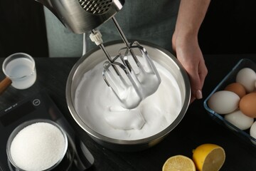 Woman making whipped cream with hand mixer at black table, closeup