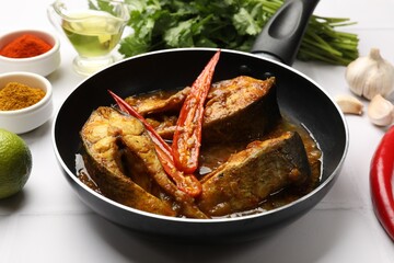 Tasty fish curry in frying pan and ingredients on white tiled table, closeup. Indian cuisine