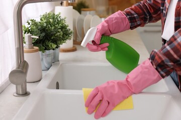 Woman with spray bottle and microfiber cloth cleaning sink in kitchen, closeup