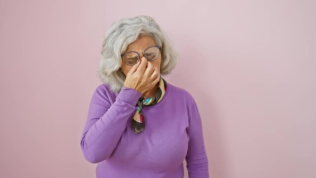 Exhausted middle-aged woman in glasses rubbing eyes, battling fatigue and stress-induced headache over pink isolated background