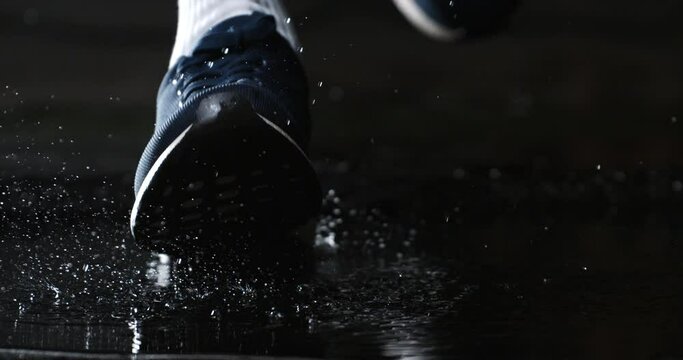 Super slow motion macro of active sportsman wearing sneakers shoes is running through puddle of rainwater with splashing drops jogging workout on street at 1000 fps.