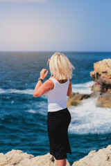 blonde in a white shirt and black skirt stands with her back and looks at the sea on a rocky shore on a sunny day