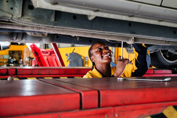 Portrait of an African female mechanic in yellow and blue uniform standing under the car bottom for inspecting in garage. Woman smiling while holding a wrench. Car repair service. Vehicle maintenance