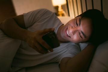 Portrait of Asian man lying in bed at night and using smartphone. Young man lying in bed using smartphone for social media and watching series at late night. Insomnia, sleep disorder concept.