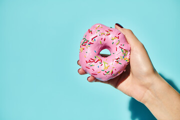 sweet delicious donut with pink frosting on it in hand of unknown female holding it on pink backdrop