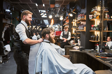 Regular client visiting favorite barbershop. Professional hairdresser and customer smiling. Male barber putting on white cape.