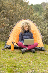 Curly and blonde hair, mountaineer woman camps with his orange tent, teleworking with his laptop