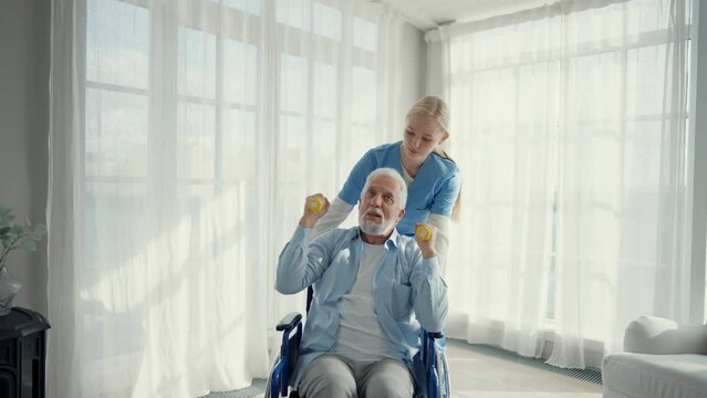 Man in wheelchair exercising with a nurse. Social medical worker woman helping senior male doing hands exercises with dumbbell. Palliative care in late stage of neurodegenerative pathologies.
