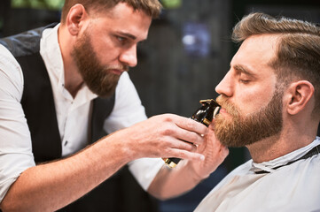 Professional hairdresser shaping thick beard. Close-up of male barber in fancy outfit shaving...