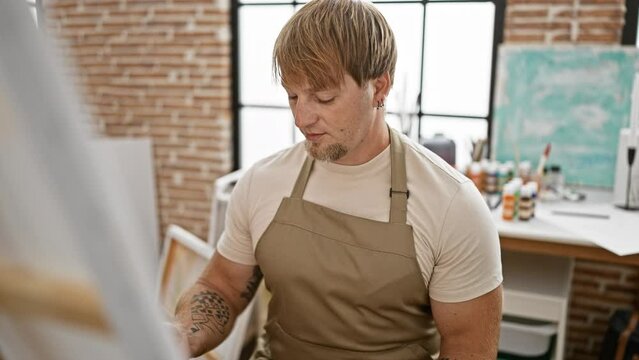 A focused, bearded, blond man wearing an apron paints in a well-lit studio with an artistic vibe.