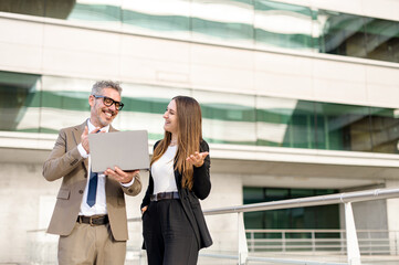 Grey-haired businessman and his female associate engage in a dynamic conversation over a laptop standing outdoors, their expressions reflecting a successful and positive exchange of ideas