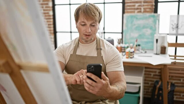A focused young man with a beard using a smartphone in a bright art studio