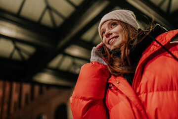 Low-angle view portrait of charming young woman in warm winter hat and jacket talking using holding mobile phone standing on dark urban street, in cold winter in night city.