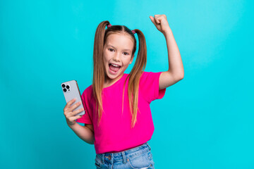 Photo of adorable overjoyed schoolgirl with ponytails hold smartphone scream yeah raising fist up...