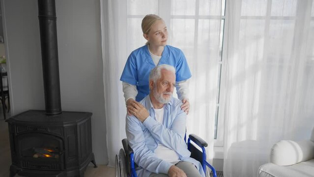 Old man in wheelchair talks with nurse. Paralyzed pensioner thanks social worker woman for help support at home. Caregiver holds hand on patient shoulder stands near. Legs paralysis, special needs.