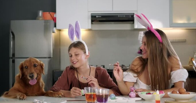 Mom and daughter wearing bunny ear hair holders sit at the kitchen table and decorate Easter eggs. Next to them is a golden retriever dog with his tongue hanging out. Family traditions.