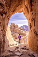 Zelfklevend Fotobehang A person in purple is walking through a natural stone archway, surrounded by rocky terrain under a clear sky, ai generative © larrui